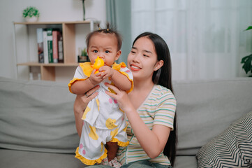 Portrait of Asian mother sits on the sofa holding her little daughter and playing with her happily in the living room at home.