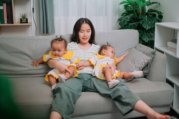 Portrait of Young mother sits on a sofa and gently holds her twin daughters in living room at home.