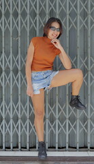 Pretty, chic Asian woman poses stylishly against a metal gate, wearing a rust crop top, distressed...