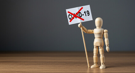 A wooden mannequin holding a sign with covid crossed out on a wooden surface against a gray background
