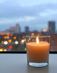Small Flame Flickers Inside Glass Cup Beside Rain-Speckled Window