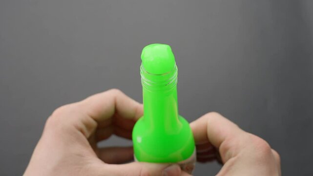 Close-up of hands pressing vibrant green floam slime through the neck of a clear bottle, creating playful texture and squishy effect, set against plain grey background, focus on tactile sensation ASMR