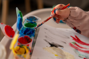 Close up of a toddlers hand reaching for a paintbrush with gouache colors. Creative activity at...