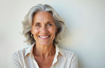 Fototapeta premium Portrait beautiful older woman smiling. Lady stands by white wall, grey hair, healthy skin, gold earrings. Mature model face expression, positive emotion. Happy old aged person, beauty in senior