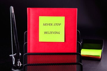 NEVER STOP BELIEVING motivational phrase on a green sticker on a red business book on a black...