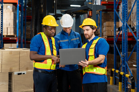 Three male warehouse workers in uniform and helmet using laptop for discussion near forklift and wrapped boxes. Collaborating with attention to detail while reviewing digital logistics data.