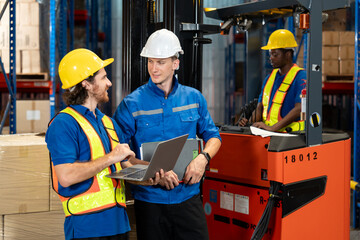 Three male warehouse employees in safety gear and uniforms working together near forklift. Two engaged in laptop discussion, third operating forklift in background. Scene inside large storage facility