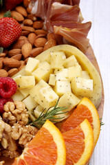 Close-up of a gourmet charcuterie and cheese board features a centerpiece of cubed cheese, possibly with herbs, surrounded by fresh almonds, walnuts, vibrant raspberries, strawberries, and oranges
