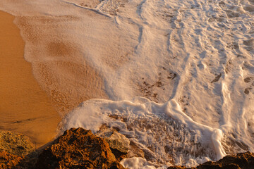 Waves wash the sandy beach in the setting sun view from above. The texture of sand and foamy waves. Beautiful natural background