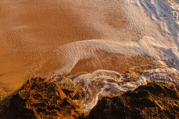 Waves wash the sandy beach in the setting sun view from above. The texture of sand and foamy waves. Beautiful natural background