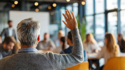 Older male learner with short gray hair raising hand during professional development training, diverse adult students seated around him, bright seminar room