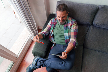 Bearded man comfortably sitting on sofa holding a tablet and a credit card, making an online...