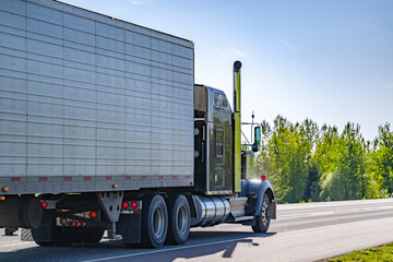 Black classic powerful big rig semi truck transporting cargo in refrigerator semi trailer running on the flat highway road