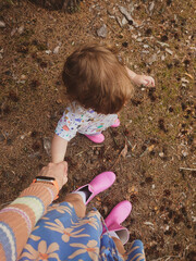 A first-person perspective shot of a walking mother and baby. The toddler is wearing pink rubber boots. The ground is covered in brown pine needles and fallen leaves. Mama is holding daughter's hand. 