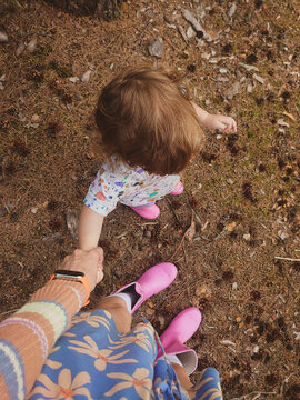 A first-person perspective shot of a walking mother and baby. The toddler is wearing pink rubber boots. The ground is covered in brown pine needles and fallen leaves. Mama is holding daughter's hand. 