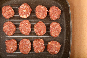 A close-up, overhead shot shows raw savory minced meat and cheese patties arranged on a dark grill pan, ready for cooking