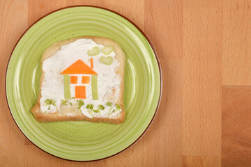 Top-down shot features a fun, edible house creation made from vegetables on a slice of bread with cream cheese, served on a green plate