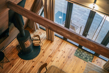 Top view of cozy Scandinavian cabin interior with modern black wood stove, wooden floor, large windows, beige curtains, and wooden terrace outside