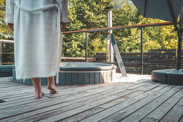 Close-up of barefoot person in white bathrobe walking on wooden terrace towards outdoor hot tub on a sunny day, relaxation and spa concept