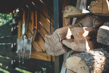 Stack of cut firewood logs stored near outdoor barbecue tools hanging on wooden wall, preparation for grilling and heating, rustic outdoor cooking area