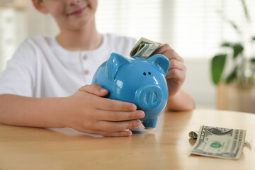 Boy putting pocket money into piggybank at table indoors, closeup