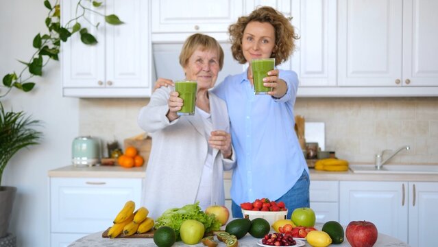 Senior woman and her daughter are showing glasses of green smoothies while standing in a kitchen full of fresh fruit and vegetables, promoting a healthy lifestyle