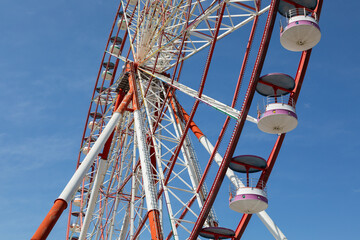 Low angle view of colorful ferris wheel with white and pink passenger gondolas and red steel structure against clear blue sky at amusement park