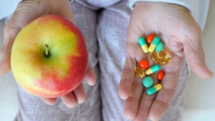 Elderly woman holds an apple in one hand and a handful of pills in the other, symbolizing the choice between natural health and pharmaceutical solutions