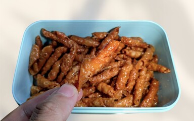 Kacang Sembunyi, an Indonesian peanut snack, fills a blue container against a white background. A hand reaches in to grab one.