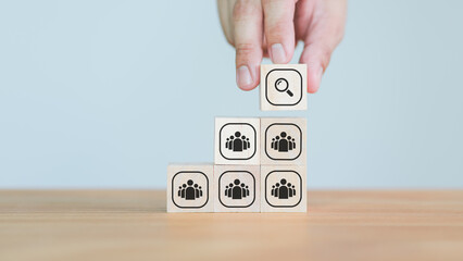 Hand placing a magnifying glass icon wooden cube block above people group wood blocks, representing talent search, company, leadership, recruitment, or leadership selection strategy.