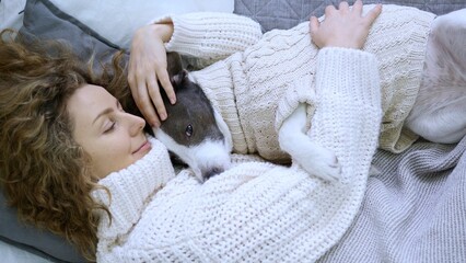 Young woman with curly hair is lying in bed with her dog, both wearing cozy knitted sweaters, enjoying a peaceful moment of affection and relaxation