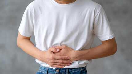 Young man holding his stomach with a painful expression wearing casual white t-shirt and jeans showing discomfort and digestive issues in indoor setting