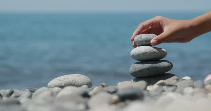Stone balancing woman creating meditative cairn on pebble beach, carefully stacking rocks with ocean waves and blue sky forming tranquil, zen like wellness scenery in background