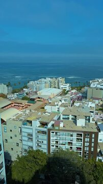High angle vertical establishing pan across coastal apartment buildings with crashing surf and turquoise waters, Lima Peru