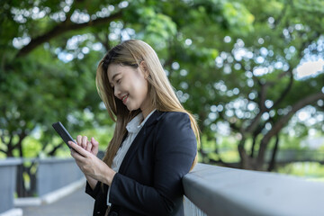 Asian Young businesswoman smiling happily while using smartphone outdoors in park with green trees...
