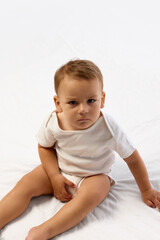 Fototapeta premium Toddler boy sits on soft bed with legs apart, frowning and holding diaper with serious face against white background. Concept of childhood, behavior, mood, emotion, independence, expression.