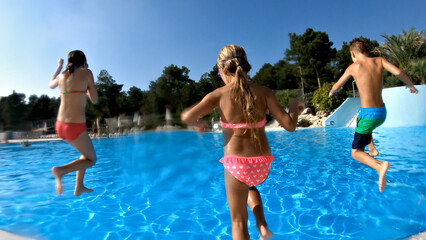 Medium shot captures three children of various ethnicities enjoying a water slide and pool at an aqua park. They wear colorful swimsuits, embodying the carefree spirit of summer fun.