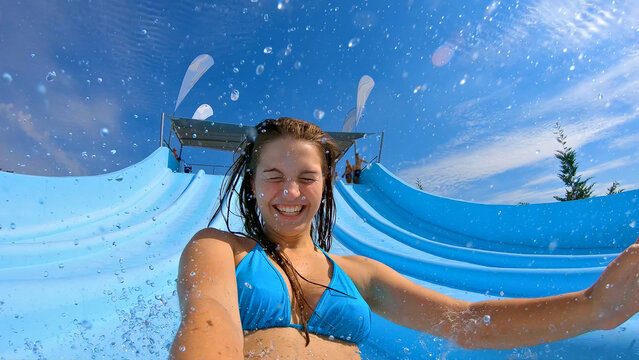 A young woman in a blue bikini splashes down a water slide, capturing the thrill and joy of a summer day at an aqua park. This is a close-up shot, emphasizing the excitement and fun of the moment.