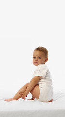 Side view photo of toddler boy sits on soft bed, looking calmly at camera over shoulder in quiet pose against white studio background. Concept of emotion, personality, childhood, kid's fashion, growth