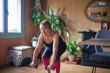 Obraz premium Senior hispanic woman smiling while working out with dumbbells at home