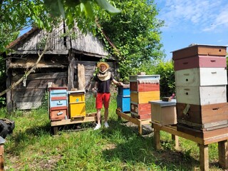 A beekeeper in protective gear stands among colorful beehives in a rustic garden, surrounded by...