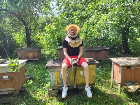 A beekeeper sits on a beehive in his protective suit, holding a smoker in the middle of a lush garden, taking a break from his work with nature and the buzzing bees