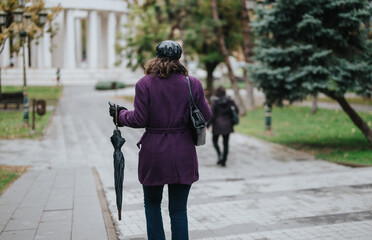 A woman dressed in a purple coat walks through a park, carrying a closed umbrella, with trees and architectural columns visible under overcast conditions.
