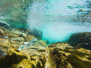 A dynamic underwater photo from a clean mountain river in Norway, capturing a powerful stream of air bubbles bursting out from between moss-covered rocks on the riverbed.