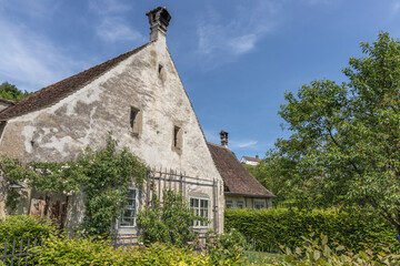 Old house with a steep, tiled roof and weathered facade, surrounded by lush greenery and trees. The white exterior shows signs of age, with patches of wear. Windows are framed with climbing plants.