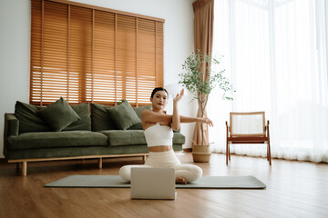 Happy young Asian woman practicing yoga and shoulder stretching at home sitting on floor in living room