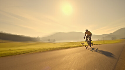 A young man of Asian descent rides his bicycle down a scenic country road, surrounded by lush green fields and mountains. The sun shines brightly, casting a warm glow on the scene. Aerial view.