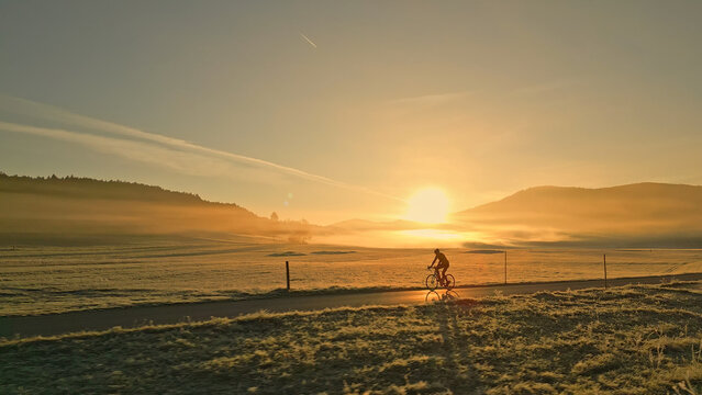 Breathtaking aerial view of a person riding a bicycle along a scenic country road during a stunning sunset. Warm hues of the sky cast a golden glow over the fields and hills.