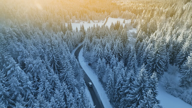 An aerial view of a winding mountain road, blanketed in snow, with a car driving through the dense, snow-covered forest. The scene evokes a sense of adventure and the thrill of alpine driving. - Powered by Adobe