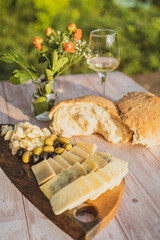 Tasty cheese and olives on a wooden board and homemade bread on the table on a sunny day in golden hour and a glass of wine and flower decoration